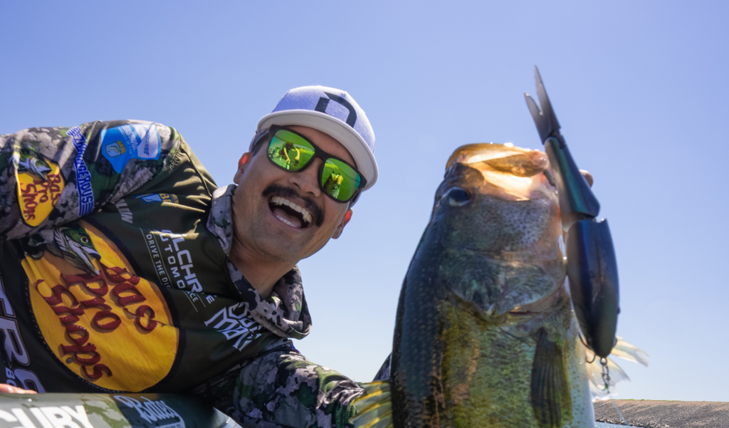 Close up of Chris Zaldain wearing an Optic Nerve cap and Optic Nerve Zaldaingerous Rumble sunglasses with a tortoise frame, amber lens, and a green mirror holding up a fish he caught with the lure in its mouth