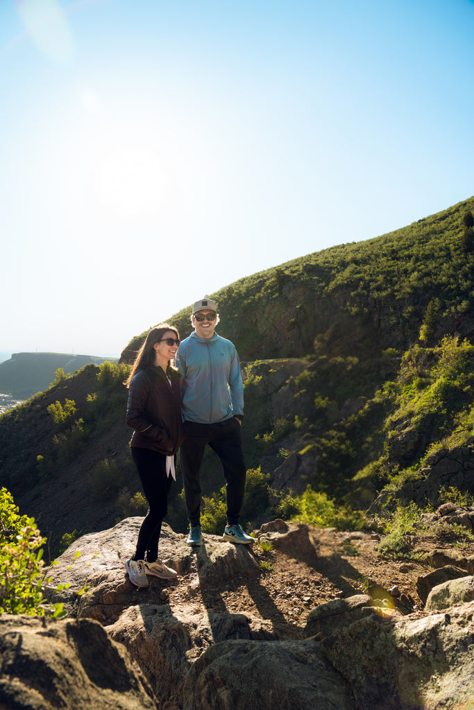 Two people wearing sunglasses standing on a rocky outcrop with a scenic background of greenery and clear sky.