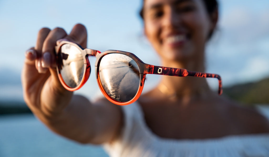 Woman holding a pair of Optic Nerve RiNO sunglasses with a tortoise frame, smoke lenses, and a gold mirror