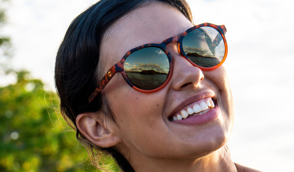 Close up of a woman wearing Optic Nerve RiNO sunglasses with a tortoise frame, smoke lenses, and a gold mirror with a blurred background