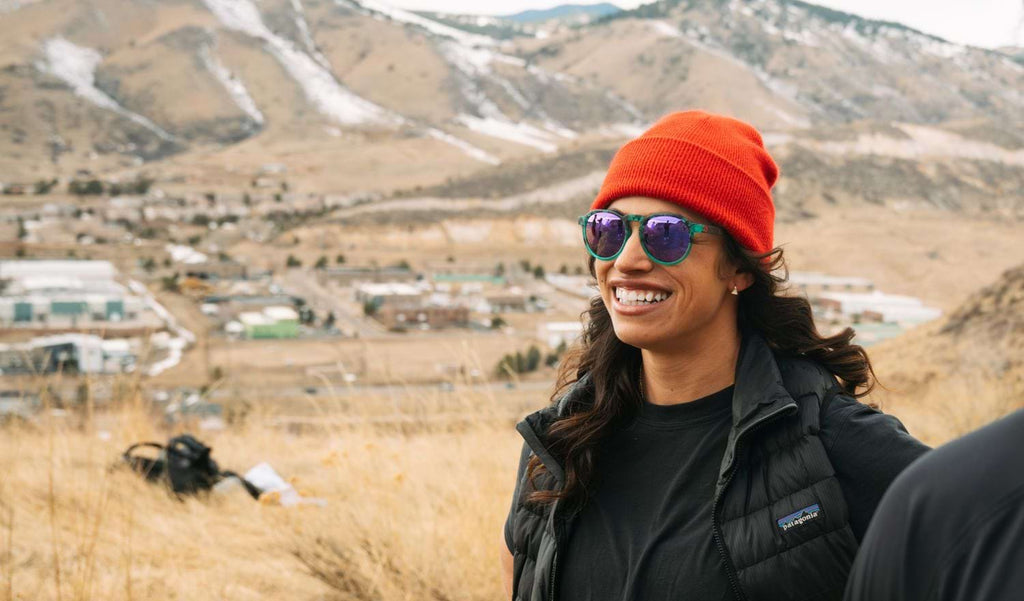 Woman wearing an orange beanie and Optic Nerve RiNO sunglasses with a blue tortoise frame, smoke lenses, and a gold mirror with mountains in the background