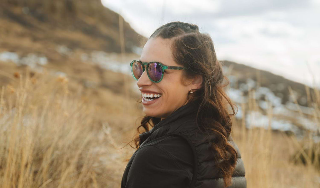 Woman wearing Optic Nerve RiNO sunglasses with a blue tortoise frame, smoke lenses, and a gold mirror with a blurred natural background.