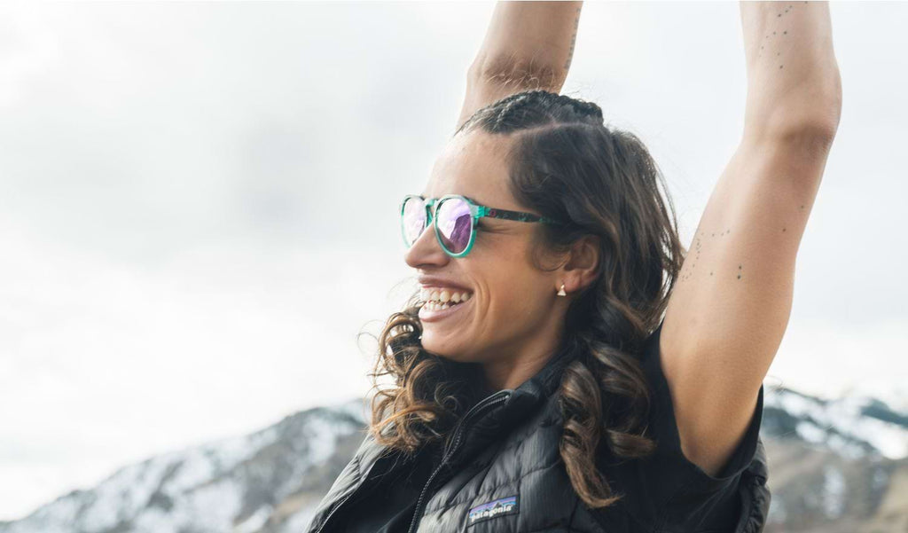 Woman with her arms above her head wearing Optic Nerve RiNO sunglasses with a blue tortoise frame, smoke lenses, and a gold mirror with a white blurred background.