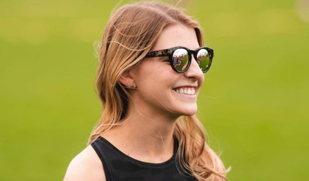 Close up of a woman wearing Optic Nerve Rizzo sunglasses with a tortoise frame, brown lenses, and a rose mirror with a blurred green background.