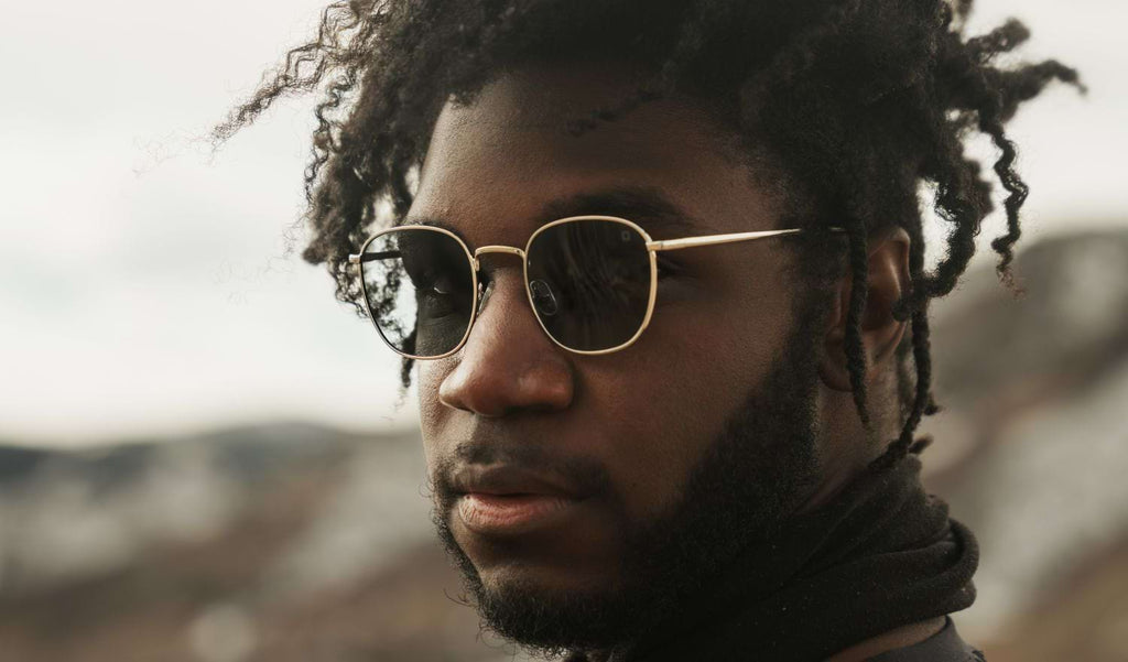 Close up of a man wearing Optic Nerve Townsend sunglasses with a gold frame and smoke lenses with a blurred background.