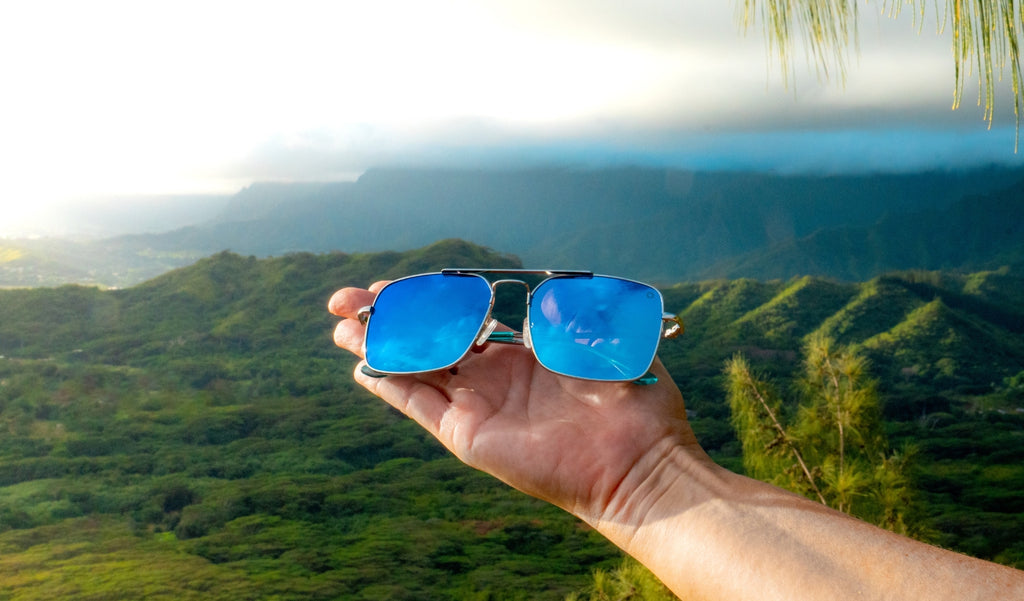 A hand holding Optic Nerve Tuner sunglasses with a silver frame, smoke lenses, and a blue mirror, with a green natural background
