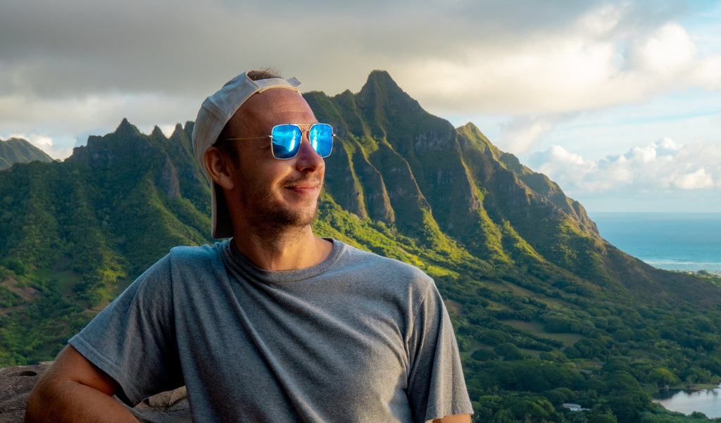 A man wearing a white baseball cap and Optic Nerve Tuner sunglasses with a silver frame, smoke lenses, and a blue mirror with a mountain in the background.