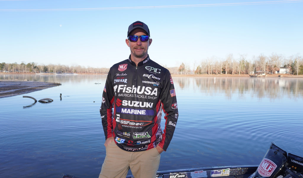 Matt Becker wearing a cap, Optic Nerve Yampa sunglasses with a black frame, brown lenses, and a blue mirror, and his professional angler jersey with a lake in the background.
