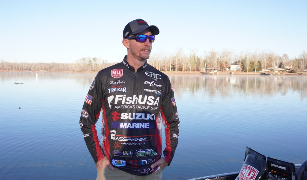 Matt Becker wearing a cap, Optic Nerve Yampa sunglasses with a black frame, brown lenses, and a blue mirror, and his professional angler jersey with a lake in the background.
