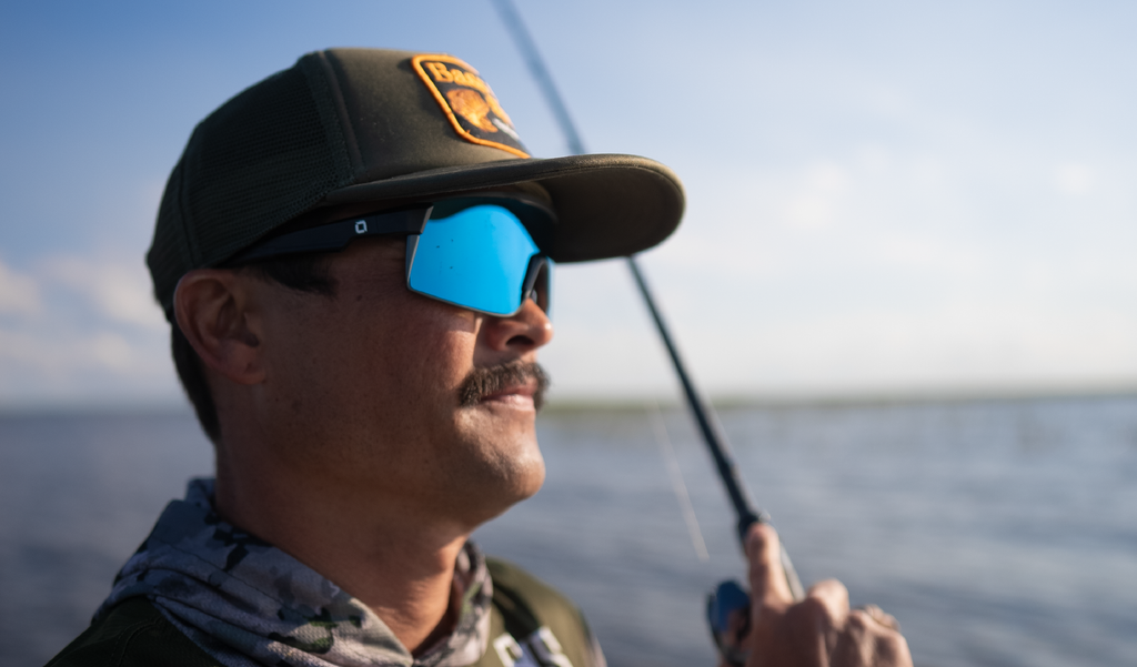 Man on a boat wearing Optic Nerve Zaldaingerous FixieMAX sunglasses with a black frame, amber lens, and a blue mirror, and a cap with a logo, holding a fishing rod.