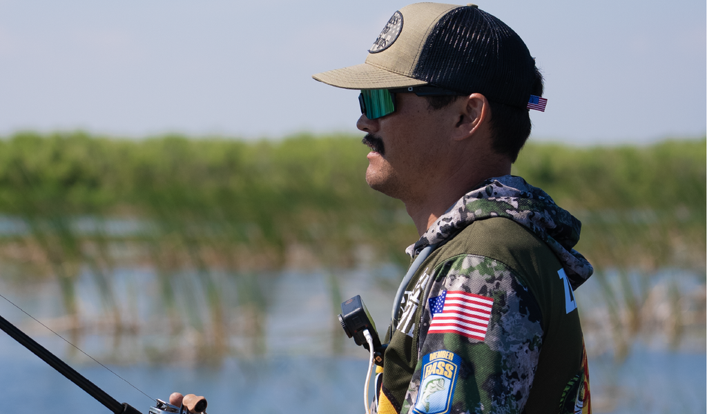 Man fishing by a body of water wearing a cap and Optic Nerve Zaldaingerous FixieMAX sunglasses with a black frame, amber lens, and a green mirror, with a blurred background