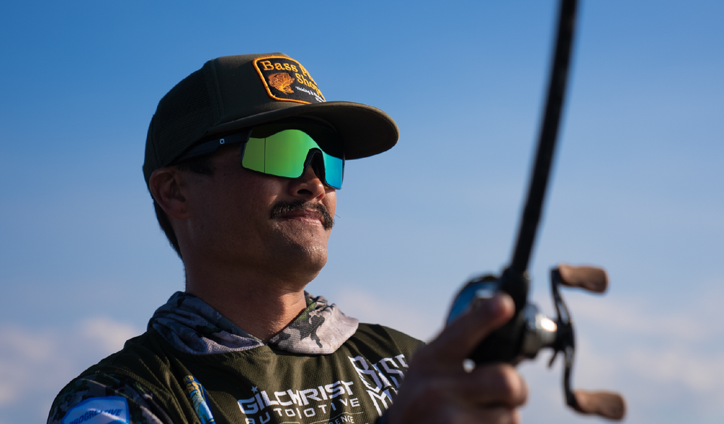 Man wearing a cap and Optic Nerve Zaldaingerous FixieMAX sunglasses with a black frame, amber lens, and green mirror holding a fishing rod against a blue sky.