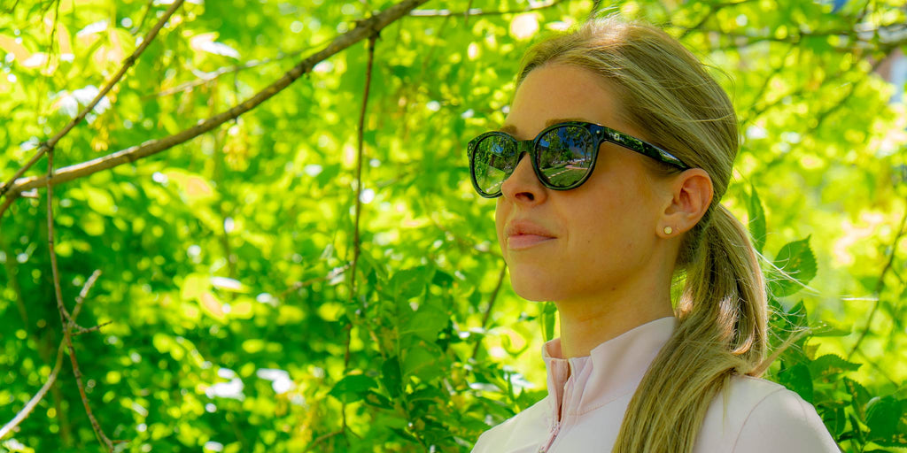 Woman wearing Optic Nerve Hotplate sunglasses with a black frame and smoke lenses, with a blurred green foliage background