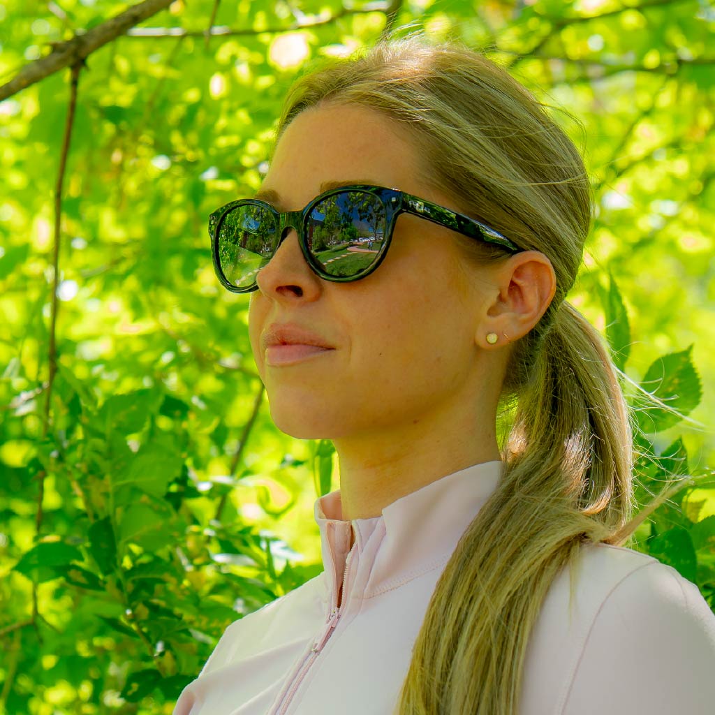 Woman wearing Optic Nerve Hotplate sunglasses with a black frame and smoke lenses, with a blurred green foliage background