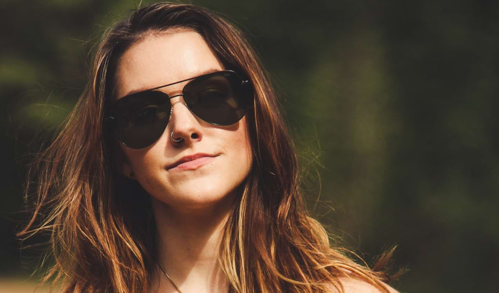 A woman wearing Optic Nerve Flatscreen sunglasses with a black frame and smoke lenses with a blurred natural background