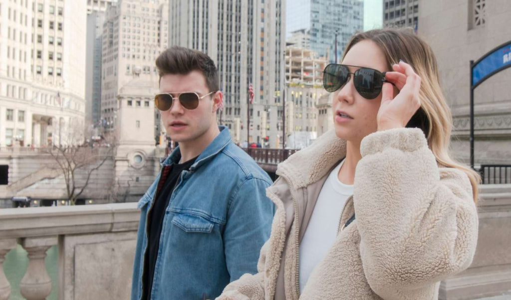 Two people walking across a bridge wearing Optic Nerve sunglasses with buildings in the background.