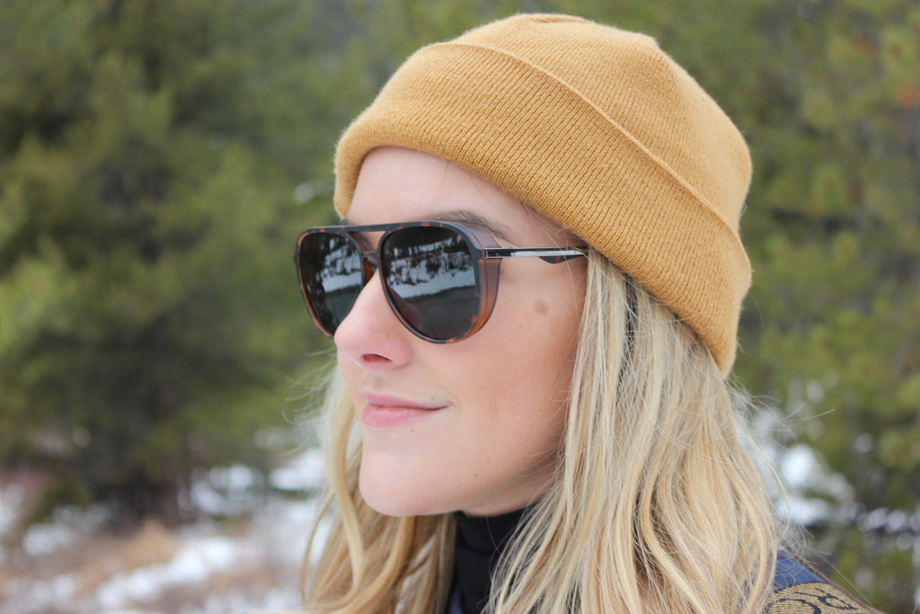 Woman wearing a yellow beanie and Optic Nerve Govnah sunglasses with a tortoise frame and smoke lenses with a blurred natural background