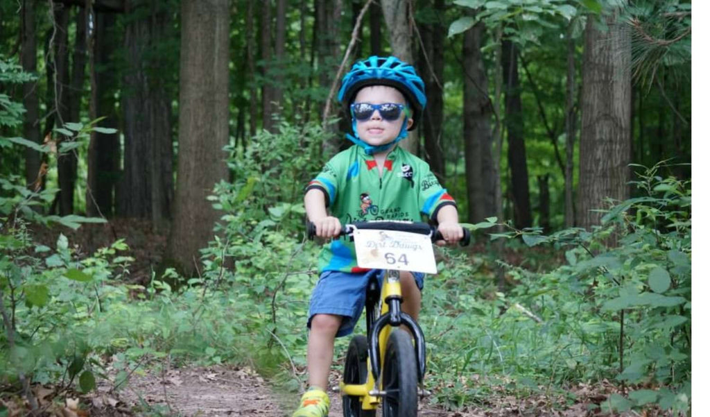 Kid riding a bicycle wearing a blue helmet and Optic Nerve Kids Boogie sunglasses with a blue frame and smoke lenses with a forest background
