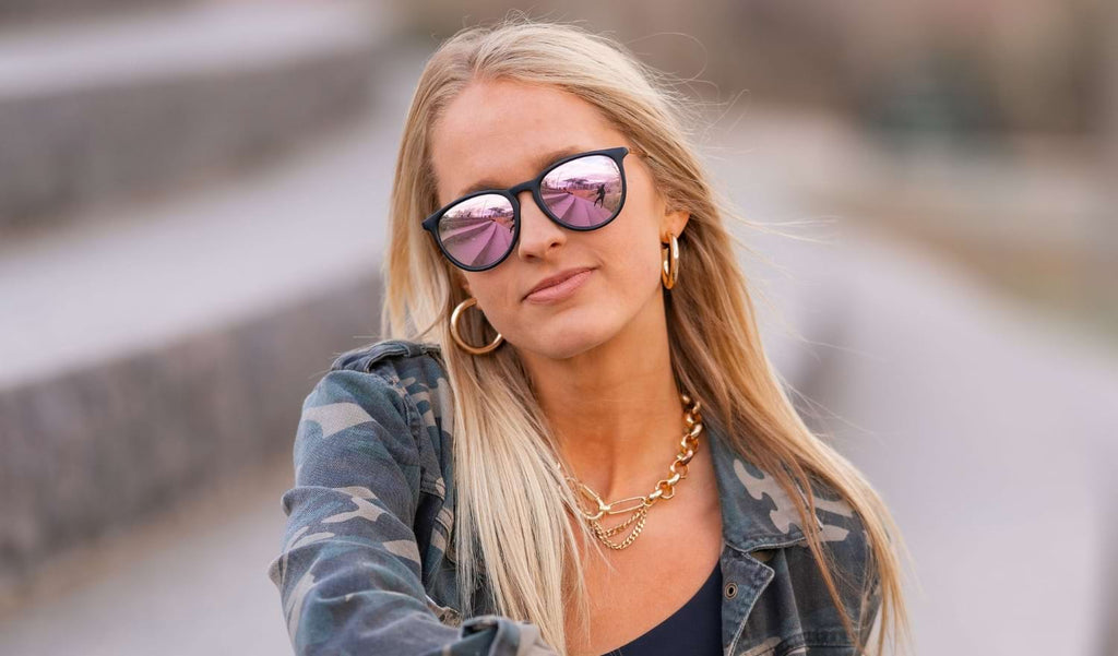 Woman wearing Optic Nerve Pizmo sunglasses with a tortoise frame, brown lenses, and a purple mirror with a blurred background