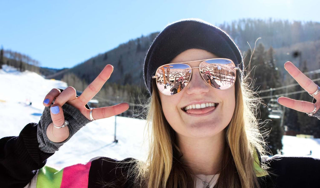 Woman wearing a black beanie and Optic Nerve Flatscreen sunglasses with rose gold frames, smoke lenses, and rose gold mirror in front of a ski lift
