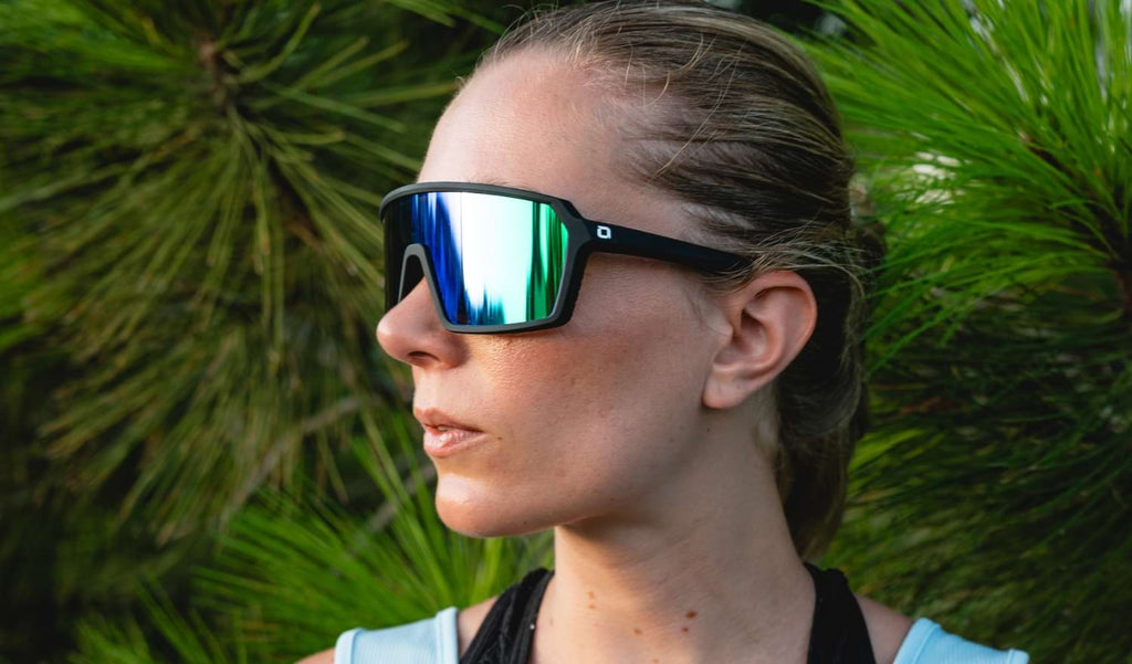 Close up of a person wearing Optic Nerve FixiePHENOM sunglasses with a black frame, smoke lens, and green mirror on a natural background
