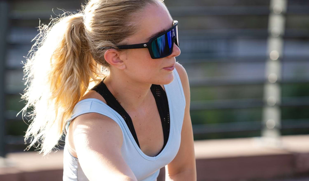 Woman wearing Optic Nerve FixiePHENOM sunglasses with a black frame, smoke lens, and a green mirror with a blurred background