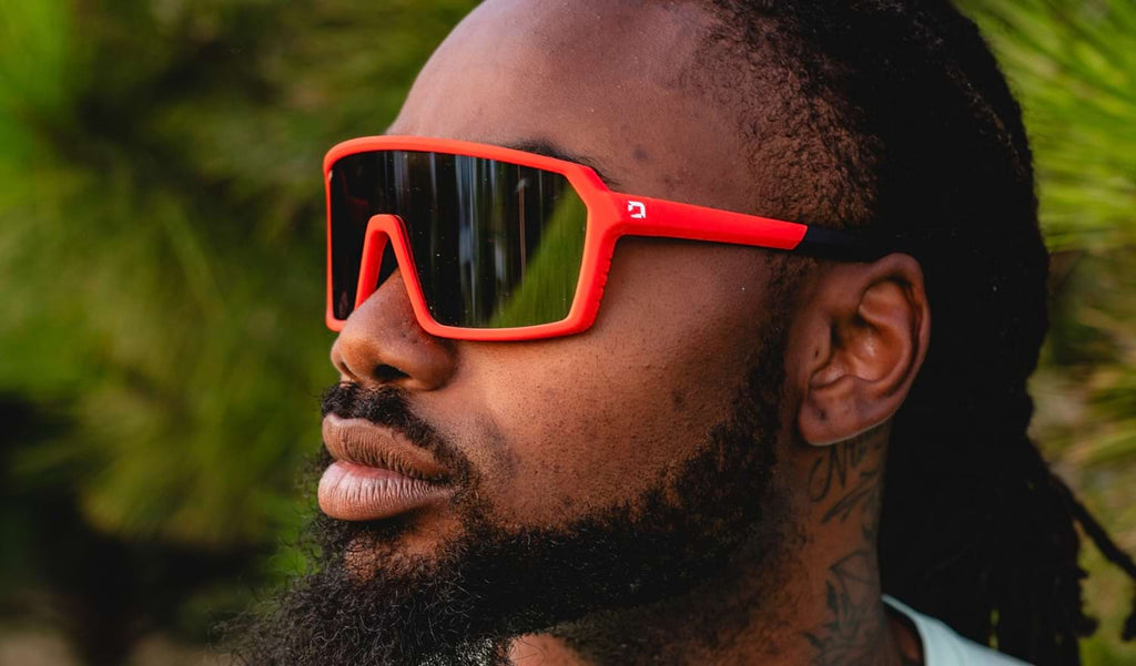 Close up of a man wearing Optic Nerve FixiePHENOM sunglasses with a red frame, brown lens, and silver mirror with a blurred natural background