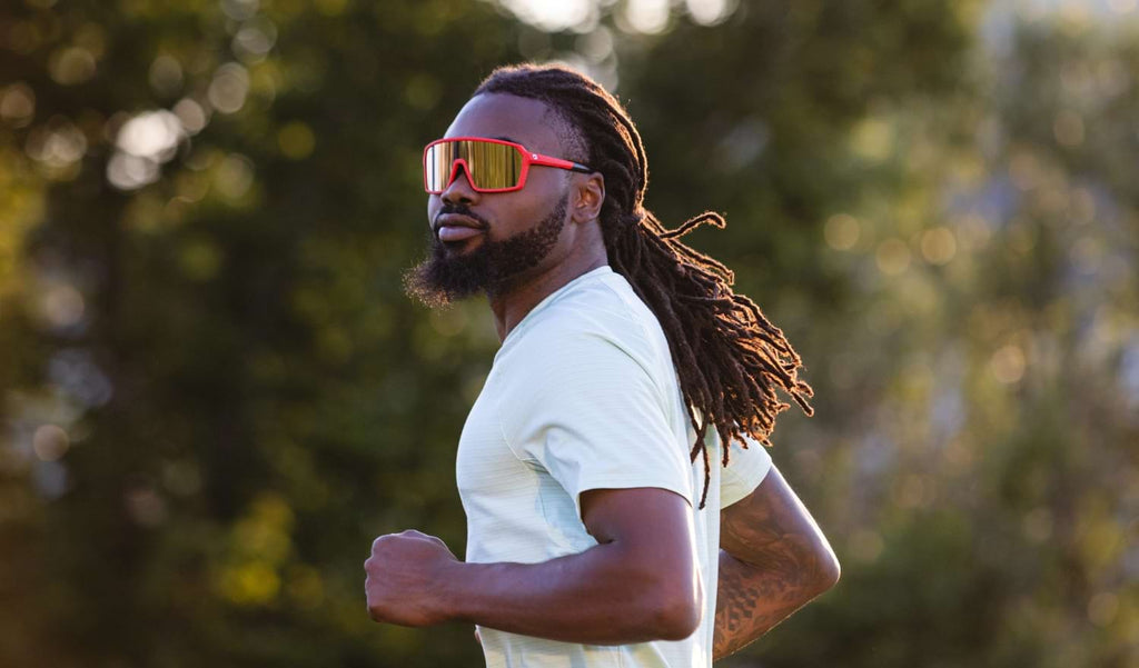 Man running wearing Optic Nerve FixiePHENOM sunglasses with a red frame, brown lens, and silver mirror with a natural blurred background