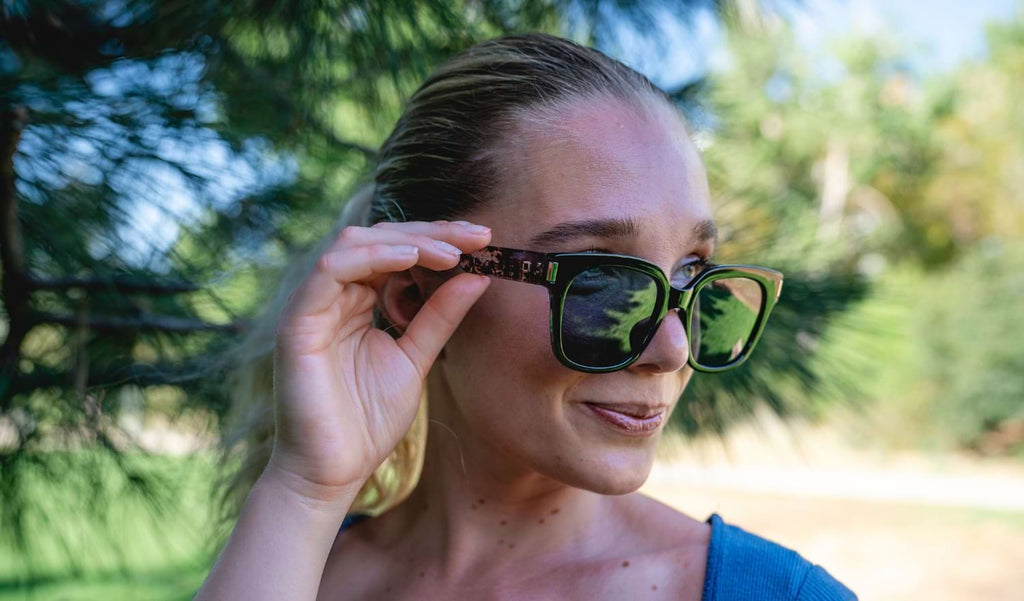 Close up of a woman wearing Optic Nerve Gracie sunglasses with a black frame and smoke lenses against a natural blurred background