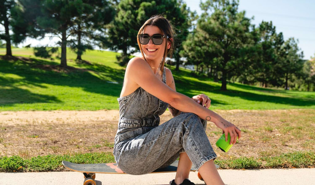 Woman sitting on a skateboard wearing Optic Nerve Gracie sunglasses with a black frame and smoke lenses holding a drink with trees in the background