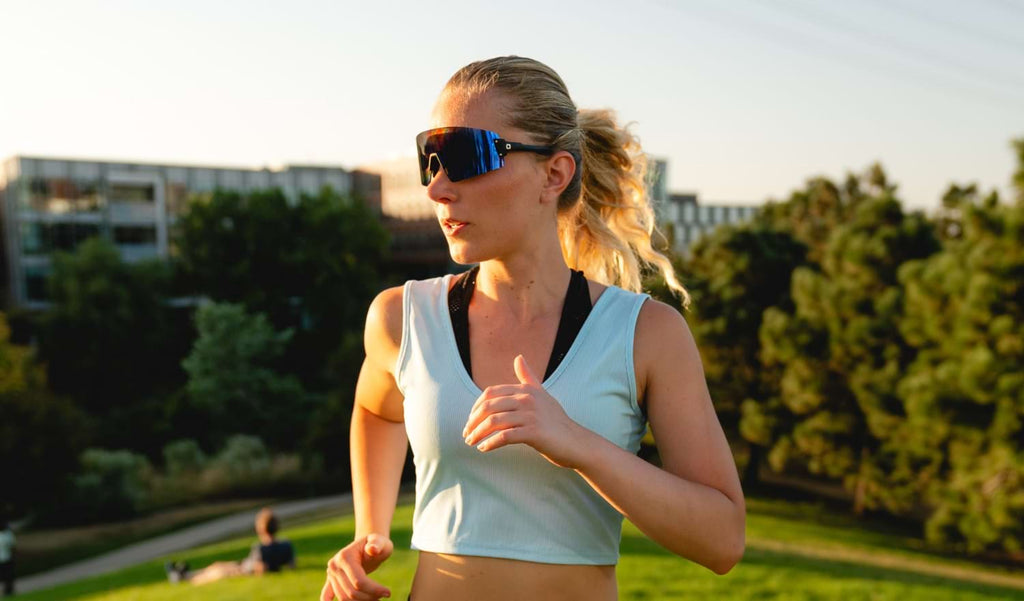 Women running wearing Optic Nerve FixieBLAST sunglasses with a black frame, smoke lens, and a blue mirror with a blurred natural background
