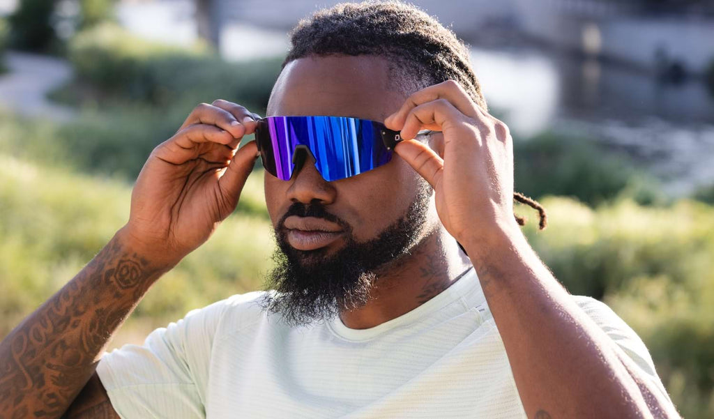 Man wearing Optic Nerve FixieBLAST sunglasses with a black frame, smoke lens, and a blue mirror with a blurred background