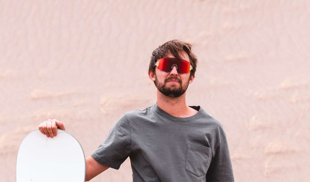 Man wearing Optic Nerve FixieBLAST sunglasses with a clear frame, smoke lens, and a red mirror with a sand dune background