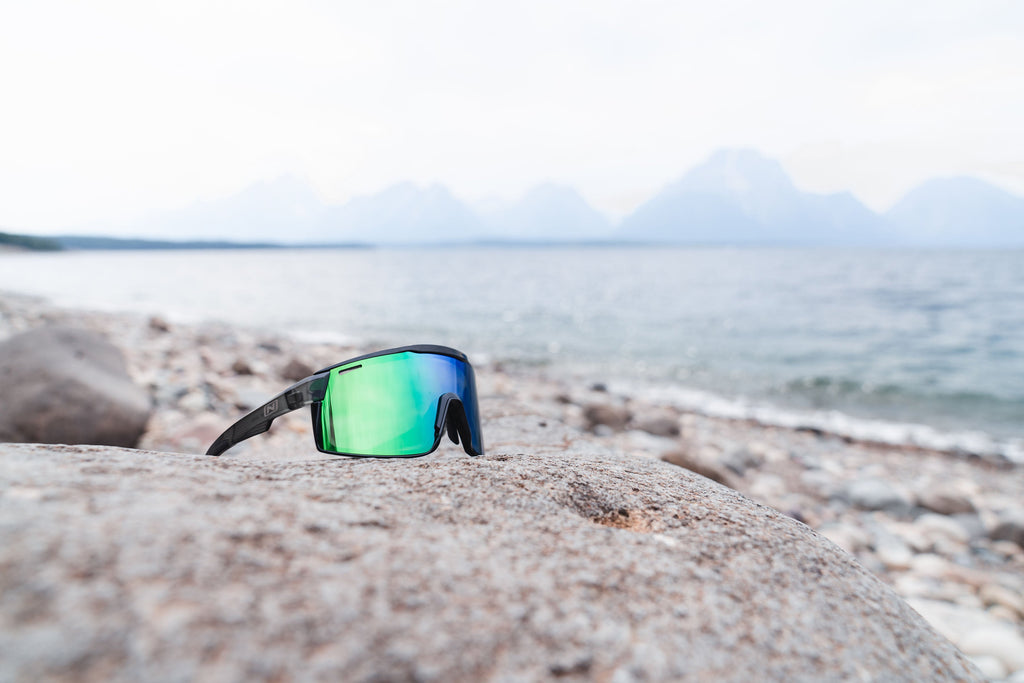 Optic Nerve FixieMAX sunglasses with a grey frame, smoke lens, and a green mirror, on a rock with the shore as its background