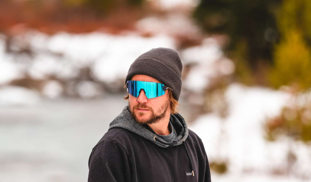 Man wearing Optic Nerve FixieBLAST sunglasses with a black frame, smoke lens, and a blue mirror with a blurred snowy background