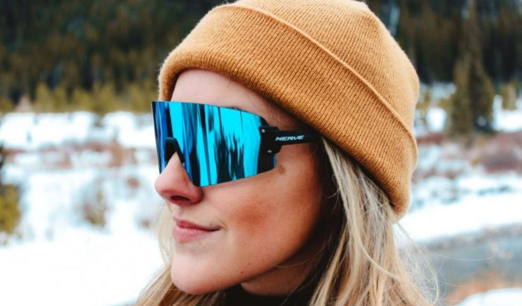 Woman wearing Optic Nerve FixieBLAST sunglasses with a black frame, smoke lens, and a blue mirror with a blurred snowy background