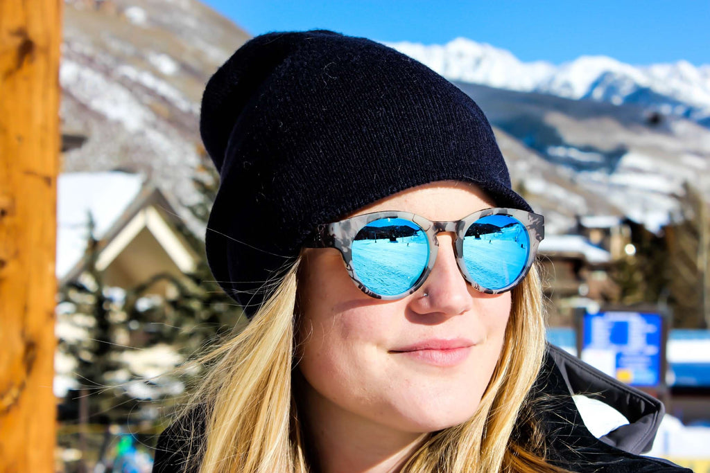 Woman wearing a black beanie and Optic Nerve Rizzo sunglasses with a tortoise frame, smoke lenses, and a blue mirror with a blurred mountainous background.