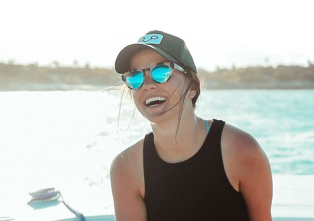 Woman on a boat wearing a cap and Optic Nerve Rizzo sunglasses with a tortoise frame, smoke lenses, and a blue mirror with a blurred lake background