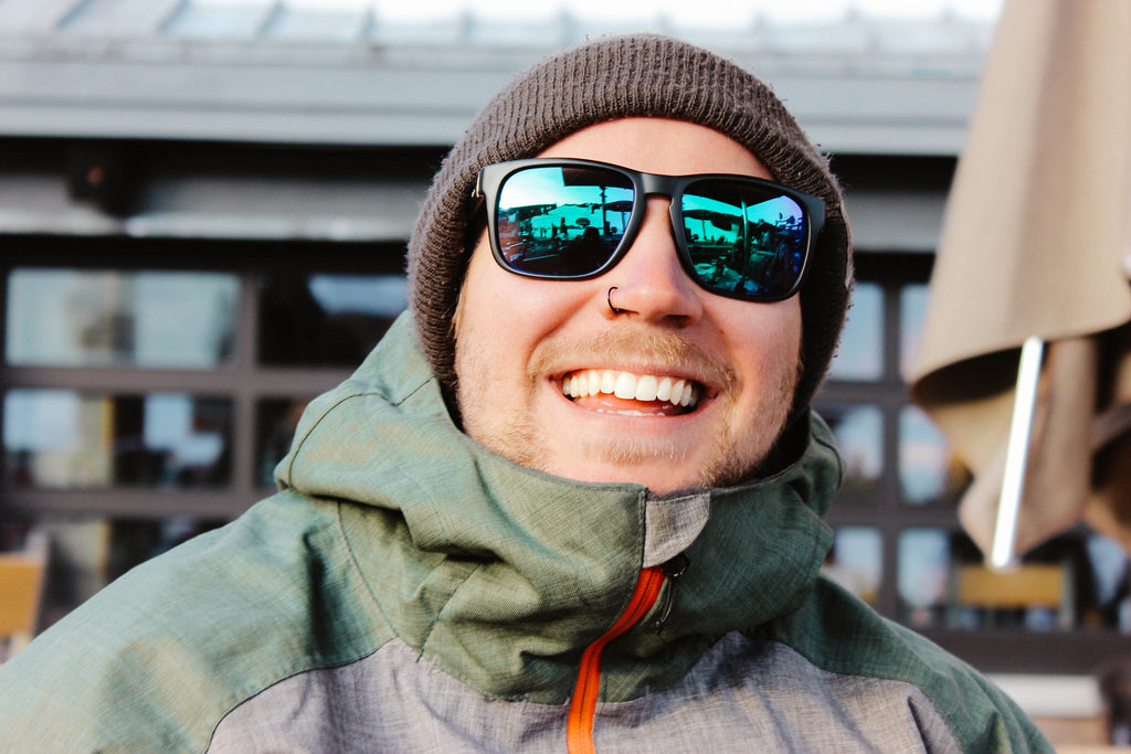 Man wearing a beanie and Optic Nerve Ziggy sunglasses with a black frame, smoke lenses, and a green mirror with a blurred background.